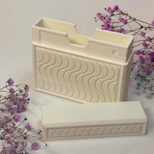 Op de afbeelding: Gebroken witte rechthoekige doos met een deksel, met een golvend patroon op de voorkant en een trapsgewijs ontwerp aan de bovenkant. Het deksel heeft een bijpassend decoratief patroon. Paarse bloemen op de achtergrond.