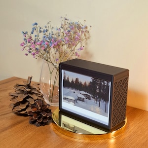 May include: A black digital photo frame showing a winter scene, on a gold circular base. The frame is next to a glass vase with dried flowers and pine cones on a wooden surface. The frame has a decorative side.
