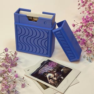 May include: A blue photo box with a wave pattern on the front, open to reveal photos inside. The lid is open, and a stack of polaroid photos is next to the box. Pink and purple flowers are in the background.
