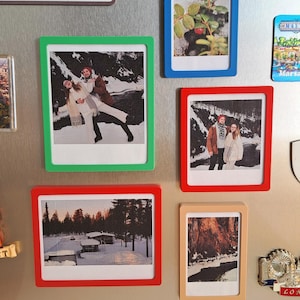 May include: A collection of framed photographs in various colors, including green, blue, red, and tan, are displayed on a metallic surface. The photos depict landscapes and people. The frames are rectangular and add a decorative element to the images.