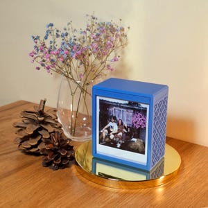 May include: A blue photo box displaying a photograph, placed on a gold-coloured tray. The box features a patterned side design. A vase of pink and blue flowers and two pine cones are also on the wooden surface.
