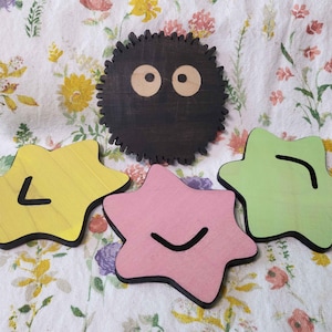 May include: Wooden coasters in the shape of stars and a round, spiky character. The star coasters are yellow, pink, and green, each with a black line design. The round coaster is black with two beige eyes.