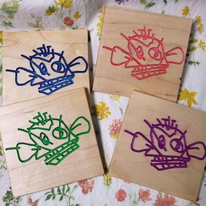 May include: Four square wooden coasters with a cartoon face design in blue, red, green, and purple. The coasters are made of light-colored wood and are arranged on a floral patterned fabric.