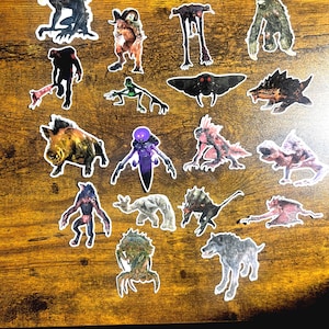 Puede incluir: Una colección de pegatinas de monstruos de varias formas y colores. Las pegatinas presentan criaturas con dientes afilados, garras y estructuras corporales únicas. Las pegatinas están dispuestas sobre una superficie de madera.
