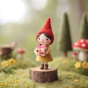 May include: A handcrafted felt gnome doll wearing a red pointed hat and yellow dress, holding a gift box with pink ribbon and heart accents. The doll stands on a wooden stump in a whimsical outdoor setting with mushrooms and trees.