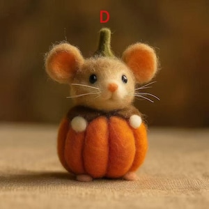 Könnte beinhalten: Eine handgefertigte Filzmaus in einem orangefarbenen Kürbiskostüm. Die Maus hat hellbraunes Fell, große orangefarbene Ohren und eine rosa Nase. Der Kürbis hat weiße Akzente und einen grünen Stiel. Eine skurrile Herbstdekoration.