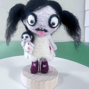 May include: A needle-felted doll with black pigtails, large white eyes, and a pink tongue, wearing a white dress with red accents. The doll holds a smaller white doll and stands on a round wooden base, supported by a thin metal rod through the center.