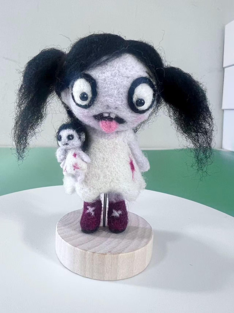 May include: A needle-felted doll with black pigtails, large white eyes, and a pink tongue, wearing a white dress with red accents. The doll holds a smaller white doll and stands on a round wooden base, supported by a thin metal rod through the center.