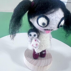 May include: A needle-felted doll with black hair in pigtails, large eyes, and a pink tongue, wearing a white dress with pink accents and dark red boots. The doll holds a smaller white doll with black hair and pink stitched details on a wooden base.