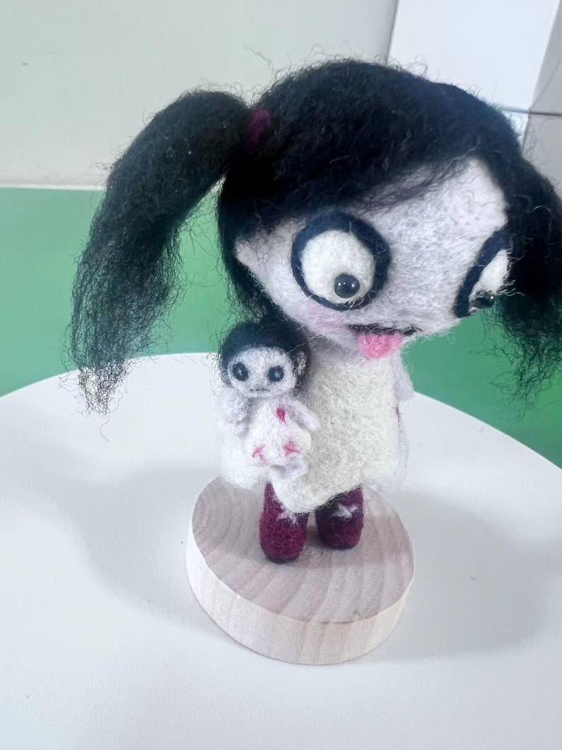 May include: A needle-felted doll with black hair in pigtails, large eyes, and a pink tongue, wearing a white dress with pink accents and dark red boots. The doll holds a smaller white doll with black hair and pink stitched details on a wooden base.