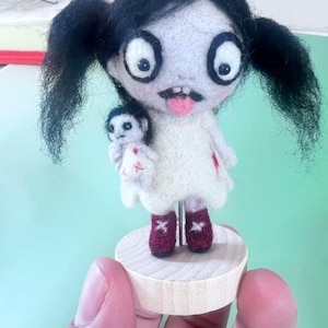 May include: A needle-felted doll with black pigtails, large white eyes, and a pink tongue sticking out. The doll wears a white dress with red accents and holds a smaller doll. It stands on a round wooden base supported by a metal rod.