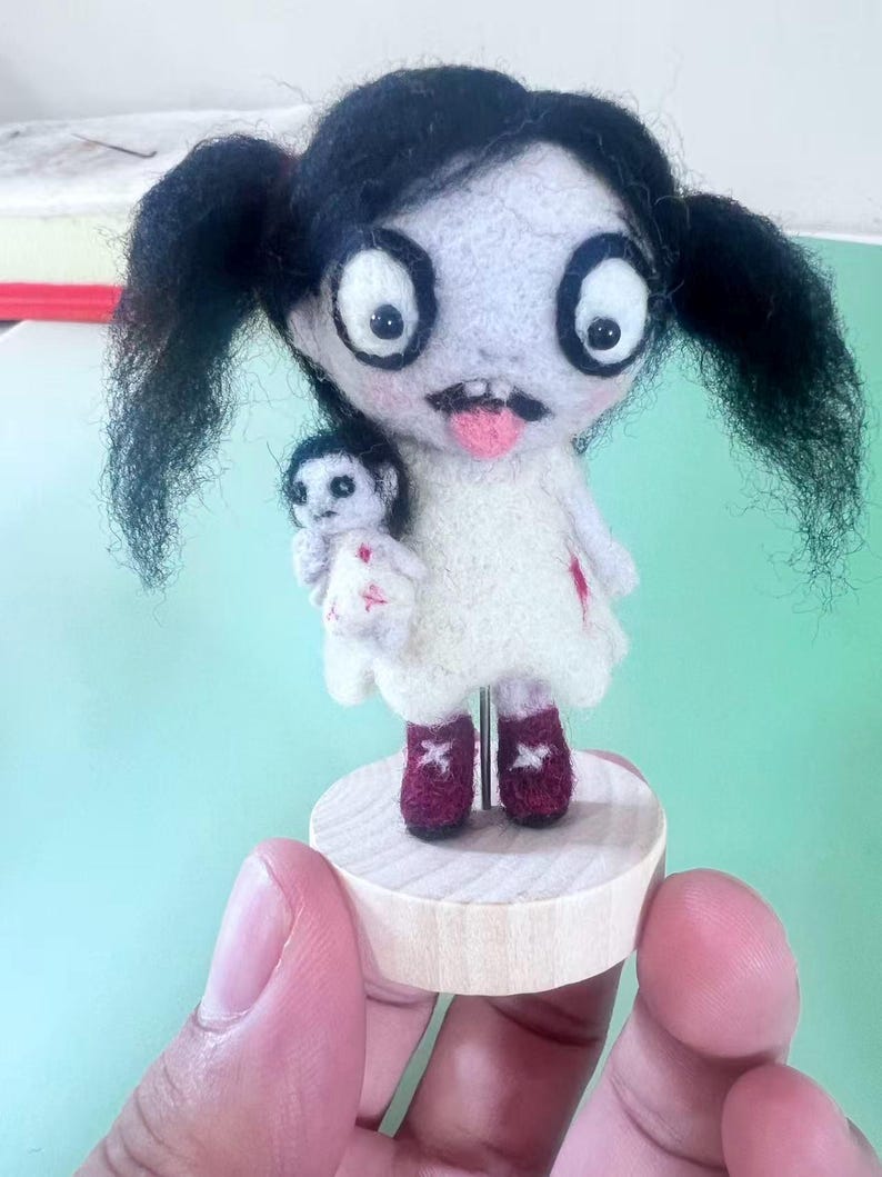 May include: A needle-felted doll with black pigtails, large white eyes, and a pink tongue sticking out. The doll wears a white dress with red accents and holds a smaller doll. It stands on a round wooden base supported by a metal rod.