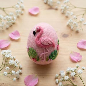 May include: A pink, egg-shaped felt ornament with a flamingo design. The flamingo is pink with a black beak, surrounded by green leaves and pink flowers. White flowers and pink petals are scattered around the ornament.