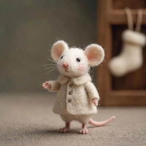 Peut inclure: Une figurine de souris blanche faite &agrave; la main, avec de grandes oreilles rondes et un nez et des joues roses. La souris porte un manteau cr&egrave;me avec trois petits boutons. Une petite queue rose s'&eacute;tend du corps de la souris. L'arri&egrave;re-plan est flou.