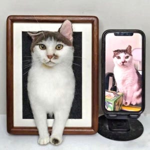 May include: A framed cat portrait with a white and brown coat. The cat is positioned in front of a black background within a wooden frame. A smartphone displays a similar image of the cat, resting on a black stand.