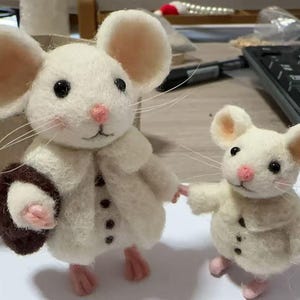 Peut inclure: Deux souris en feutre faites &agrave; la main, une grande et une petite, se tiennent c&ocirc;te &agrave; c&ocirc;te. La plus grande souris porte un manteau cr&egrave;me avec des boutons noirs et un sac &agrave; main marron. Les deux souris ont des nez, des oreilles et des pieds roses. La plus petite souris tient la main de la plus grande.