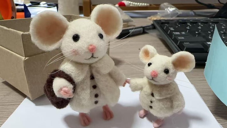 Peut inclure: Deux souris en feutre faites &agrave; la main, une grande et une petite, se tiennent c&ocirc;te &agrave; c&ocirc;te. La plus grande souris porte un manteau cr&egrave;me avec des boutons noirs et un sac &agrave; main marron. Les deux souris ont des nez, des oreilles et des pieds roses. La plus petite souris tient la main de la plus grande.