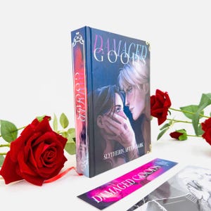 Peut inclure: Un livre à couverture rigide intitulé "Damaged Goods" avec une couverture bleu foncé représentant deux personnages. Un marque-page avec le même titre et une illustration en noir et blanc sont également présents. Des roses rouges sont disposées autour du livre.