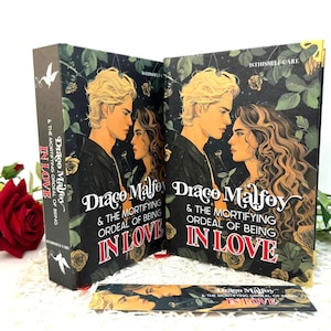 Könnte beinhalten: Zwei Hardcover-Bücher und ein Lesezeichen mit dem Titel "Draco Malfoy & The Mortifying Ordeal of Being in Love". Die Cover zeigen ein Paar vor einem dunklen Hintergrund aus Rosen und Blättern. Der Buchrücken lautet "In Love".