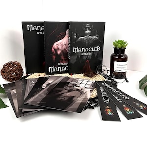 May include: A selection of books and promotional items. The books are black with the title "Manacled" in white, and feature illustrations. There are also black and white prints and bookmarks with colourful stained-glass designs.