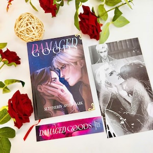 May include: A book titled "DAMAGED GOODS" with a cover illustration of two people looking at each other. A bookmark and a print with similar artwork are also present. Red roses and a decorative ball surround the books.