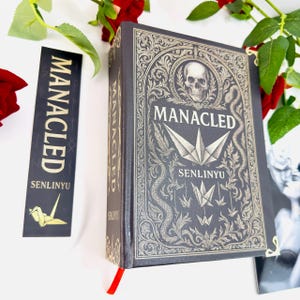Op de afbeelding: Een donkergrijs boek met de titel "MANACLED" in gouden letters, met een schedel- en origami-ontwerp. Een bijpassende bladwijzer met dezelfde titel en een gouden origami-kraanvogel is ook aanwezig. Rode rozen en groen omlijsten het boek.