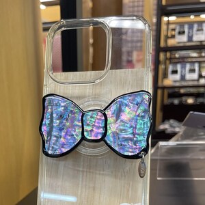 May include: Clear iPhone 14 Pro case with a decorative iridescent bow design. The bow has a black outline and a shimmering, multi-colored surface. A small silver charm hangs from the case. The case is transparent, allowing the phone's color to show through.