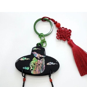 May include: Black keychain featuring a stylized mask design with iridescent accents, a bird, and cloud details. The keychain has a green ring, red tassel, and decorative beads. A unique accessory.