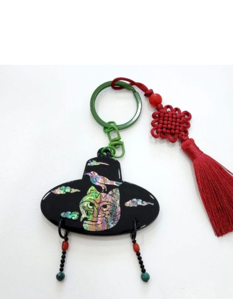 May include: Black keychain featuring a stylized mask design with iridescent accents, a bird, and cloud details. The keychain has a green ring, red tassel, and decorative beads. A unique accessory.