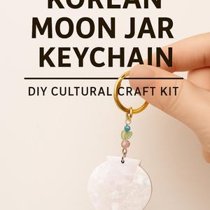 May include: A DIY Korean Moon Jar keychain kit. The keychain features a white, pearlescent, moon jar pendant with a gold-colored ring and colorful beads. Text at the top reads "MAKE YOUR OWN KOREAN MOON JAR KEYCHAIN".