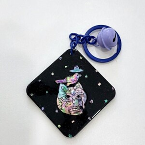 May include: A black, diamond-shaped keychain with a colorful cat face design and a bird wearing a hat. The keychain has a blue ring and a purple bell attached.