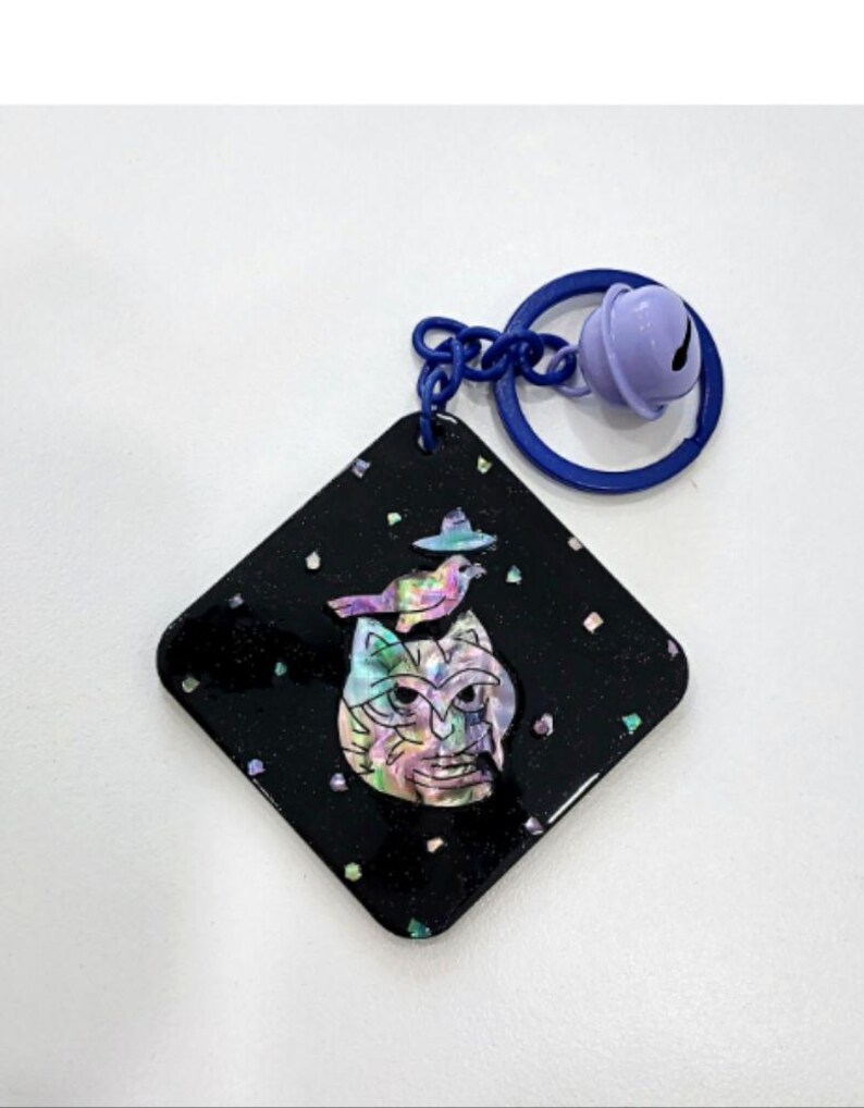 May include: A black, diamond-shaped keychain with a colorful cat face design and a bird wearing a hat. The keychain has a blue ring and a purple bell attached.