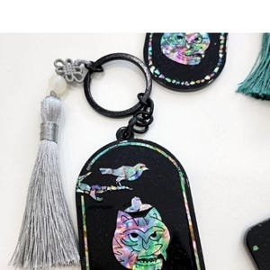 May include: Black keychain with a decorative pendant featuring a colorful, iridescent cat face design and a bird. The keychain has a silver tassel and a black metal ring. The pendant is framed with a colorful border.