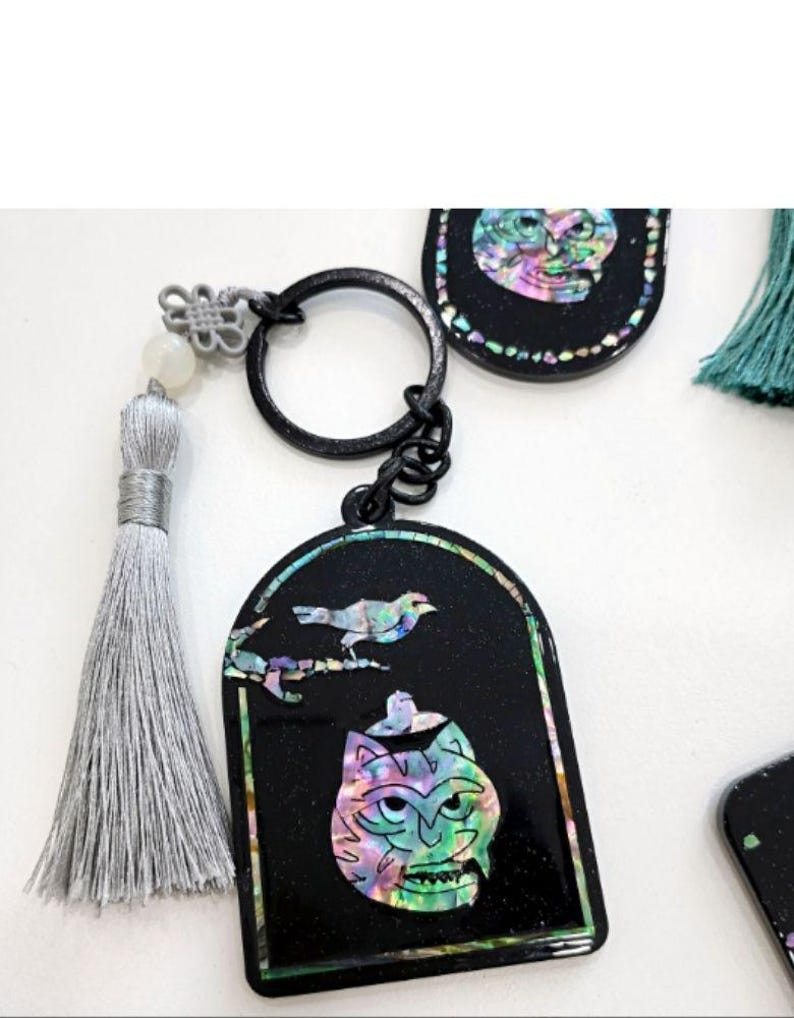 May include: Black keychain with a decorative pendant featuring a colorful, iridescent cat face design and a bird. The keychain has a silver tassel and a black metal ring. The pendant is framed with a colorful border.