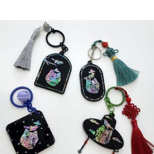 May include: A collection of black keychains with iridescent cat designs. Each keychain has a different shape, including a square, oval, and arched design. Some keychains have tassels in various colors, including gray, teal, and red. The keychains are connected to key rings.