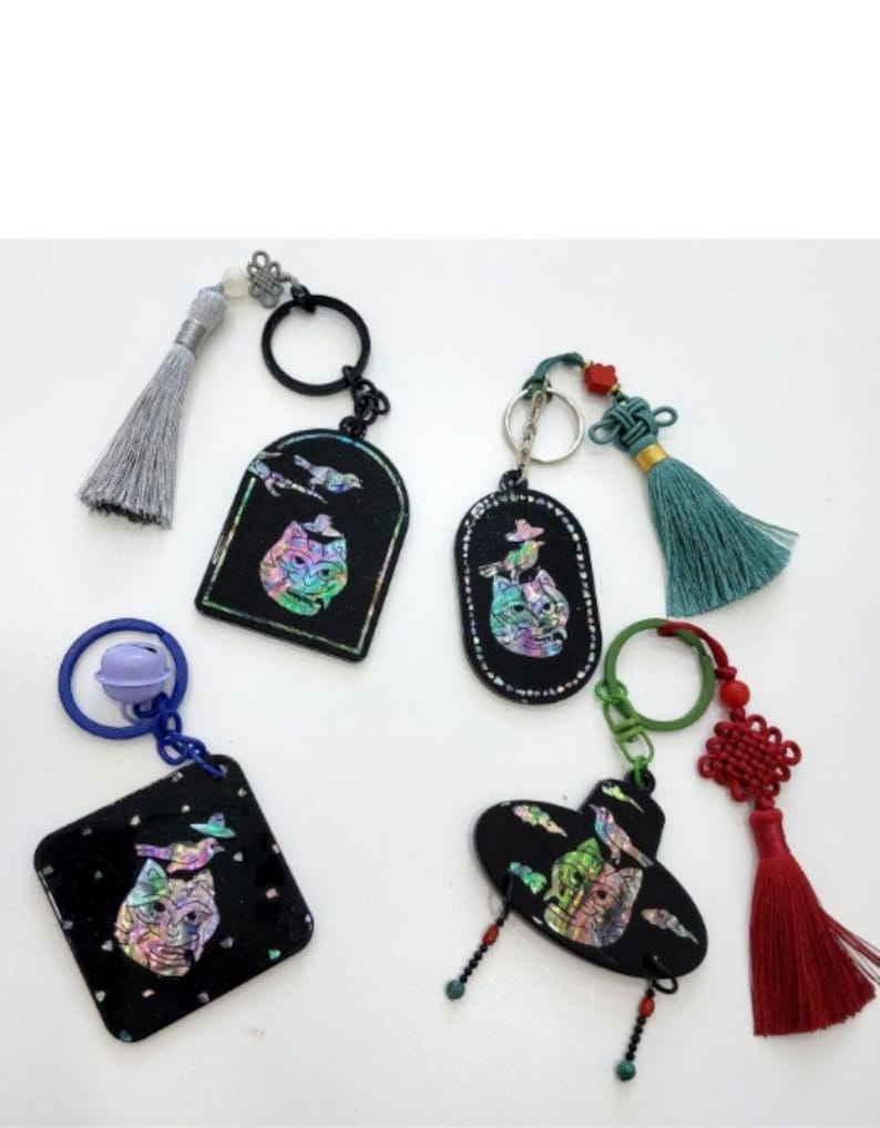 May include: A collection of black keychains with iridescent cat designs. Each keychain has a different shape, including a square, oval, and arched design. Some keychains have tassels in various colors, including gray, teal, and red. The keychains are connected to key rings.