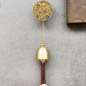 May include: A gold-colored wind chime with a circular top featuring a detailed dragon design. A gold chain connects the top to a bell-shaped chime and a wooden tag with Korean text. The chime hangs against a gray background.