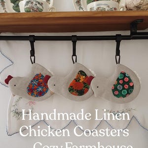 May include: Three handmade linen chicken coasters in a cozy farmhouse style. Each coaster features a gray body, a red comb, and a patterned design on the body. The text on the image reads "Handmade Linen Chicken Coasters - Cozy Farmhouse Style."