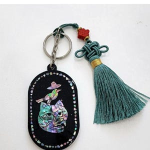 May include: Black oval keychain with a cat and bird design. The cat's face is iridescent, and the bird is perched on its head. The keychain has a silver chain and a teal tassel with a red accent.