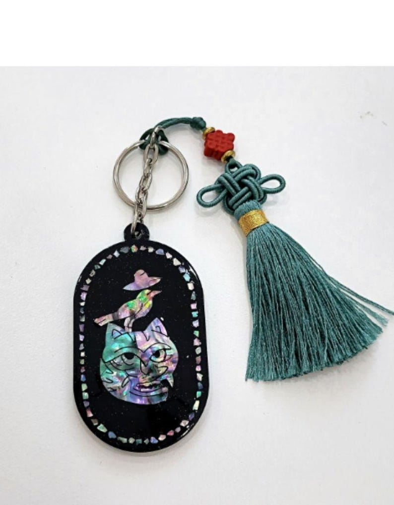 May include: Black oval keychain with a cat and bird design. The cat's face is iridescent, and the bird is perched on its head. The keychain has a silver chain and a teal tassel with a red accent.