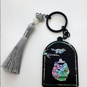 May include: Black keychain with a decorative pendant featuring a bird and a cat-like face with iridescent accents. The keychain includes a gray tassel and a black ring for attaching keys.