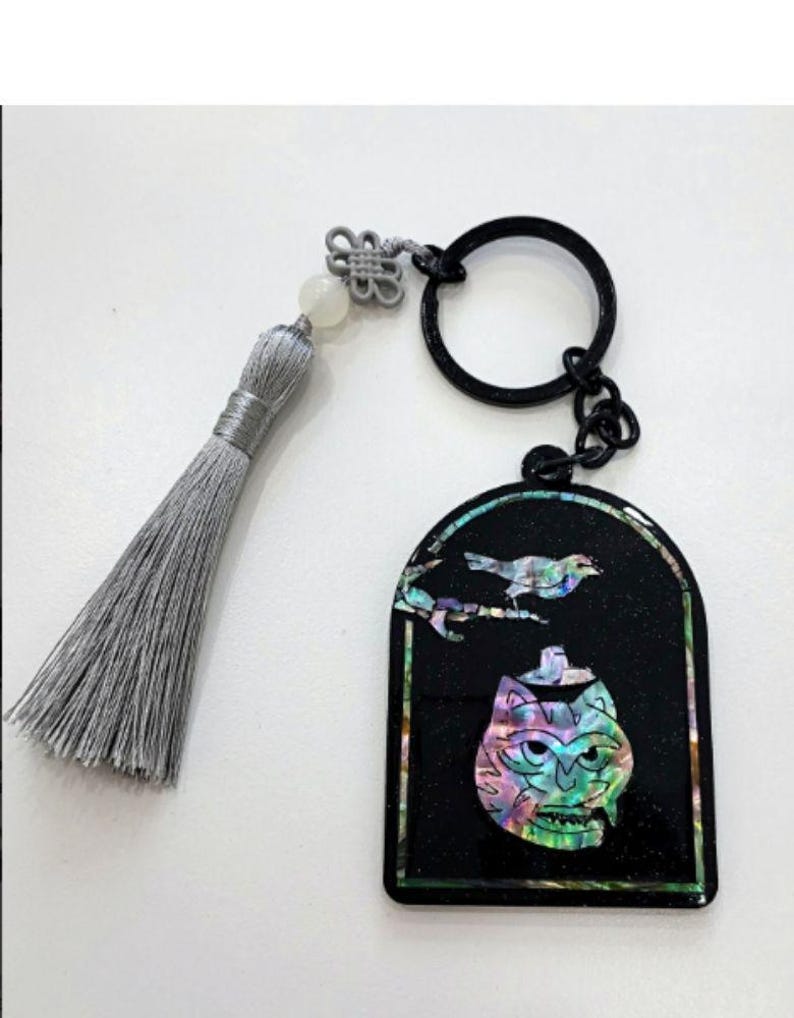 May include: Black keychain with a decorative pendant featuring a bird and a cat-like face with iridescent accents. The keychain includes a gray tassel and a black ring for attaching keys.
