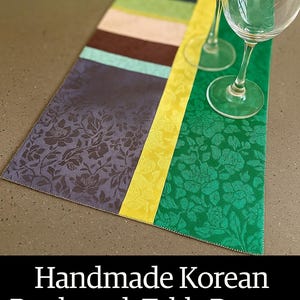 Op de afbeelding: Handgemaakte Koreaanse patchwork tafelloper met een Hanbok-geïnspireerd ontwerp. De loper heeft een kleurblokontwerp met bloempatronen in groen, paars en geel. Twee wijnglazen zijn erop geplaatst.