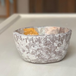 May include: A small, round, hand-formed bowl with a rough, textured exterior featuring a mottled white and earthy brown finish. The rim is decorated with a series of evenly spaced vertical notches, and the interior reveals patches of yellow.