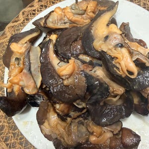 May include: A white plate piled high with cooked, dark brown, and tan-colored shellfish. The pieces appear to be sliced, showing a mix of dark, firm outer edges and lighter, fleshy, beige-colored interiors with a glossy, moist texture throughout.