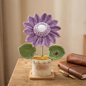 May include: A handmade crochet flower in a pot. The purple flower has a white center and green leaves with a small red ladybug. The pot is beige with a wooden base. The label reads "HANDMADE Limited edition."