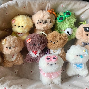 May include: A collection of handmade plush dog keychains in a woven basket. The dogs are various colors, including yellow, brown, and white, and are adorned with accessories like sunglasses, bows, and bibs. The basket is lined with a floral fabric.