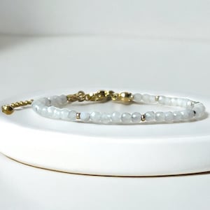Könnte beinhalten: Zartes Armband mit kleinen, facettierten, hellblau-grauen Perlen. Das Armband hat goldfarbene Metallakzente und einen Verschluss. Der Schmuck wird auf einer weißen Oberfläche präsentiert.
