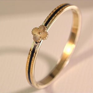 Könnte beinhalten: Goldfarbenes Armreif-Armband mit einem schwarzen Streifen und einem vierblättrigen Kleeblatt-Anhänger. Das Armband hat ein strukturiertes Design an den Rändern und eine polierte Oberfläche. Der Kleeblatt-Anhänger ist mittig auf dem Armband.