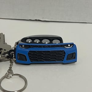May include: A blue and black car-shaped keyring. The keyring features a detailed front end design with a black grill, headlights, and a silver top. The keyring is attached to a key ring and a key.